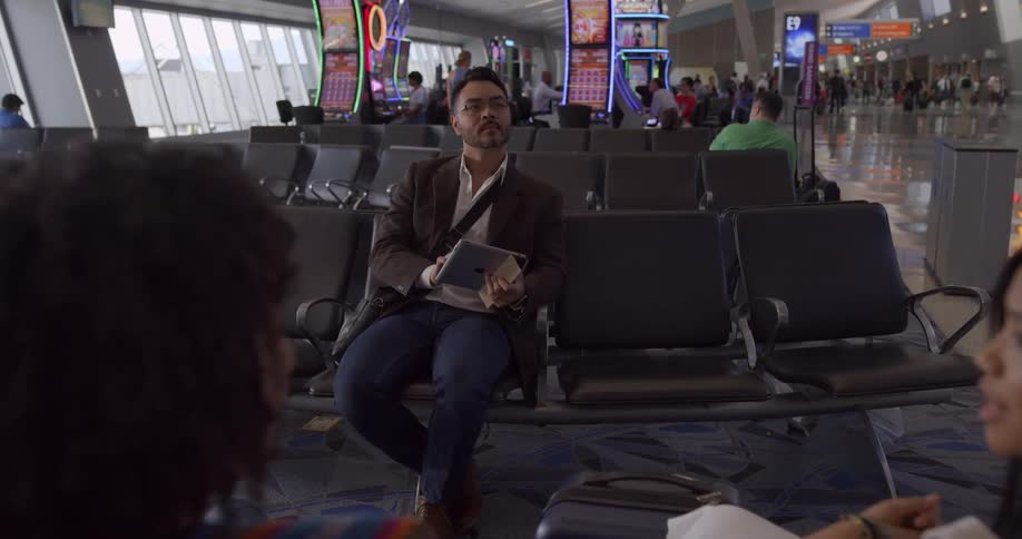 Two women sit and chat at an airport terminal waiting area while a man sits across from them reading something off of his tablet