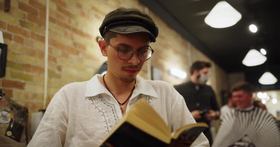 A young man wearing glasses and a hat reading a book sitting in a barber shop