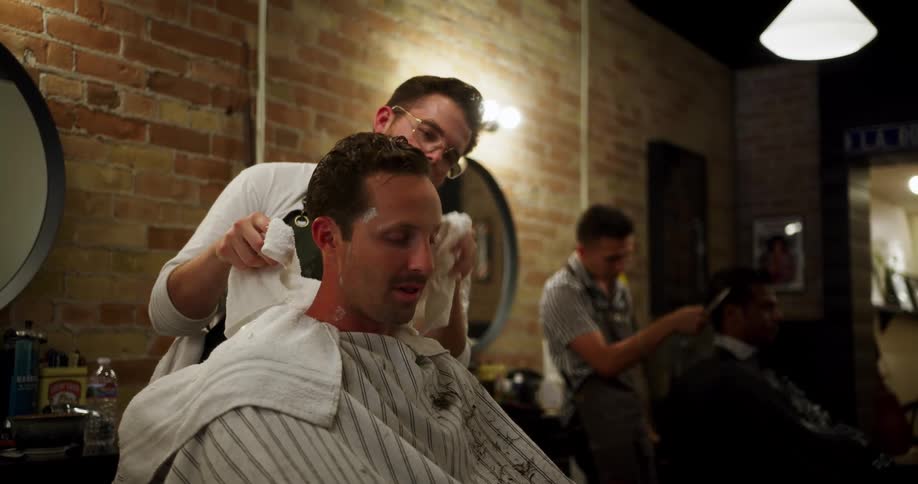 A male barber uses a towel to clean his clients head off of shaving cream