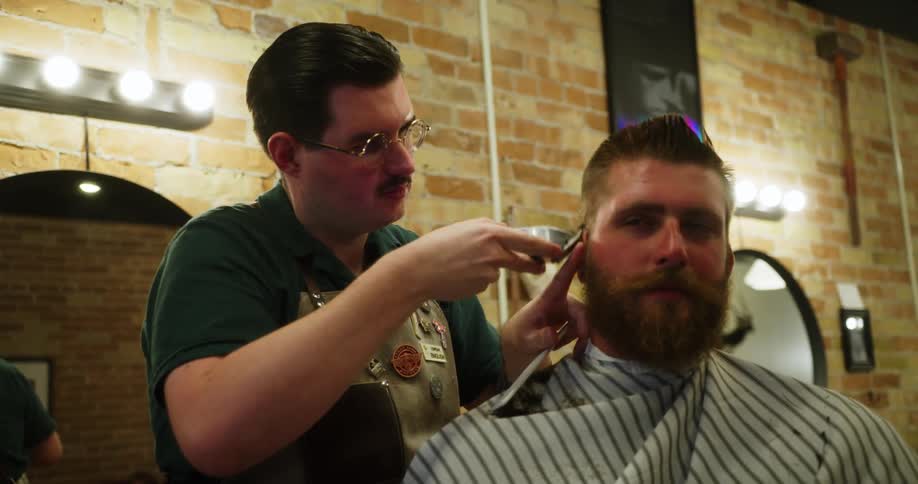 A barber carefully trims the hair of a bearded male customer