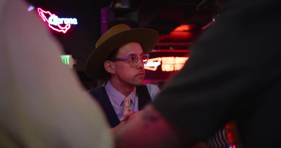 Man wearing a hat and glasses talks to friends in a bar