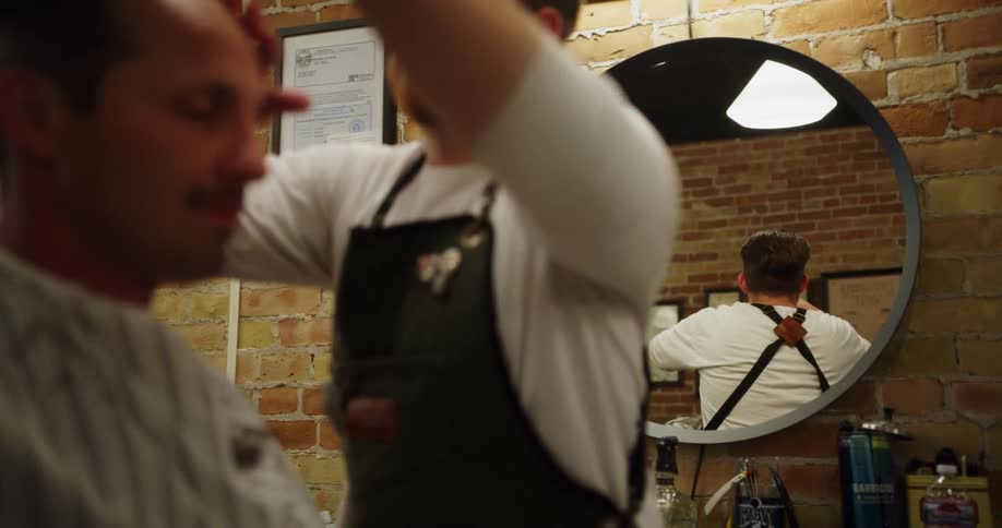 Barber moves around man in barber chair cutting their hair in front of round mirror hung on brick wall