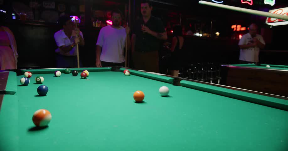 A group of people in a bar stand around a pool table as a man in a cowboy hat starts to play