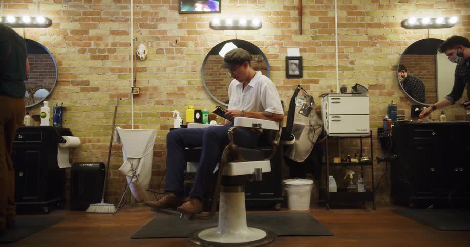 A man takes a seat in a styling chair in a barber shop and opens his book