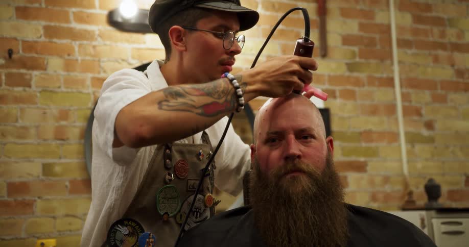 A barber closely shaves a bearded mans bald head