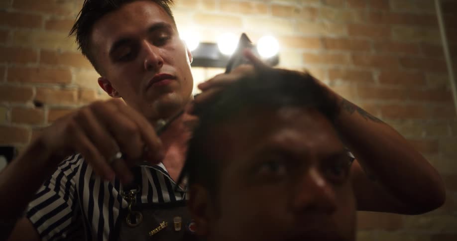A man sits in a barbers chair as his hair is combed and cut by another man in front of a brick wall with a round mirror