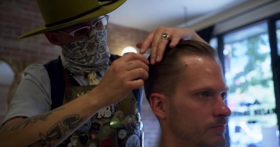 Barber wearing a hat with glasses and a bandana mask combs a mans hair