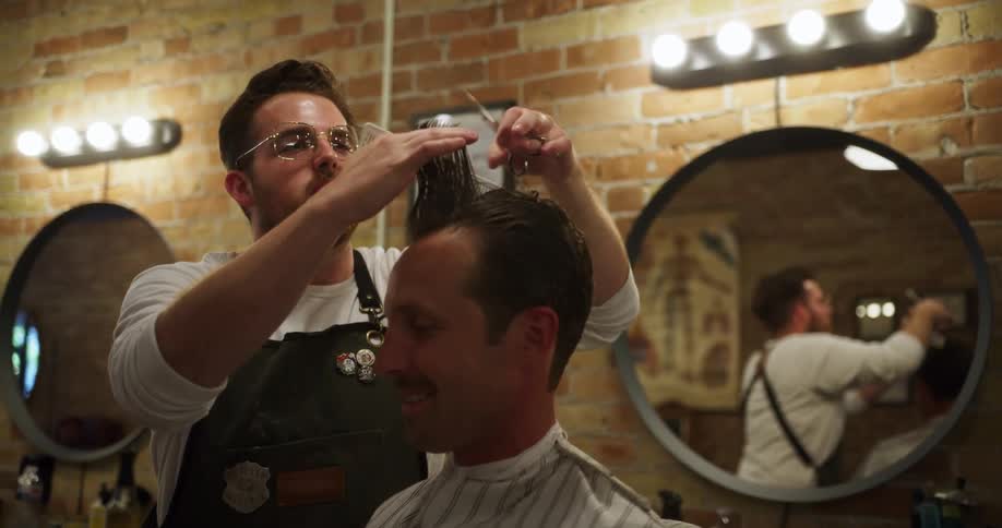 Barber wearing glasses combs a mans hair and cuts it with scissors in hand