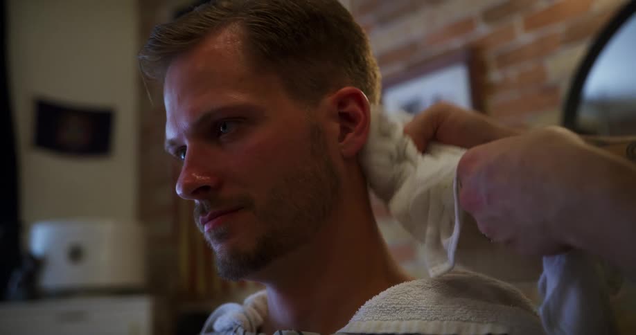 A barber wearing a mask wipes hair off a mans face with a towel