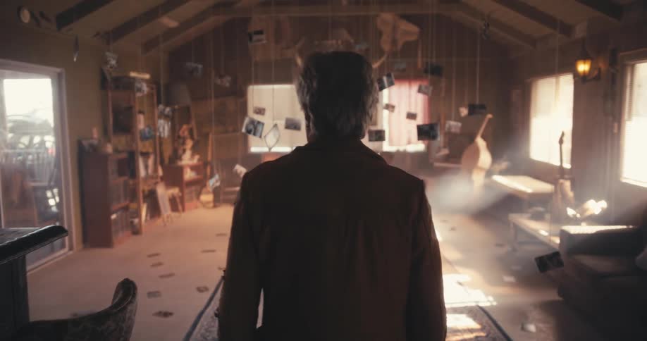 A man walks through a room with musical instruments and furniture with photos hanging from strings from the ceiling and sun streaming through windows