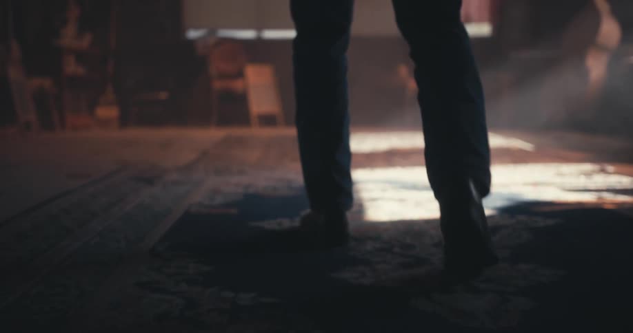 A person wearing jeans and boots walks in a room on a wood floor covered with a rug with sunlight coming through a window