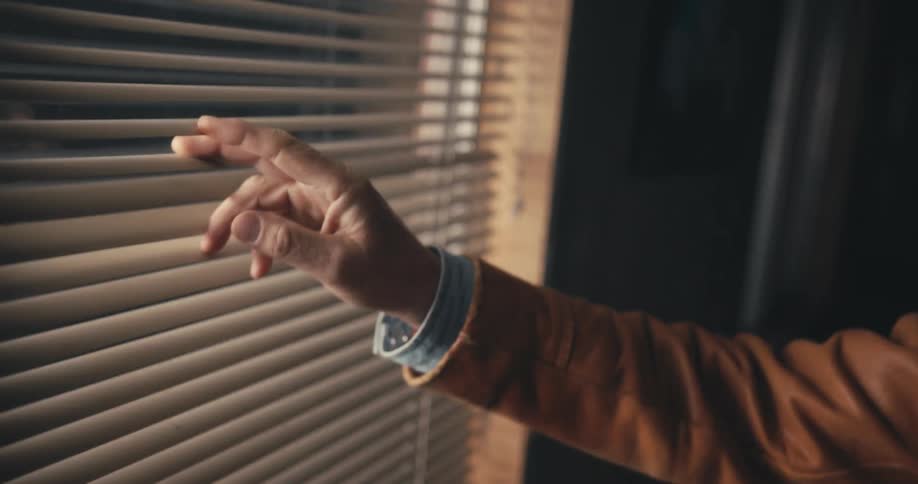 A hand pushes up the blinds slightly to look outside before slowly dropping their hand away
