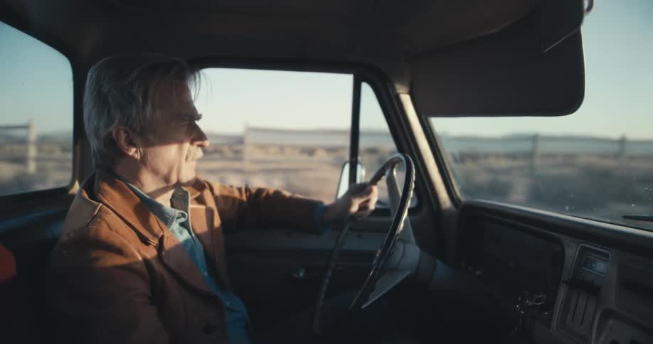 Man drives into the sunset in a truck down a road in the desert he sighs while driving he is wearing a brown jacket and denim shirt he has one hand on the wheel