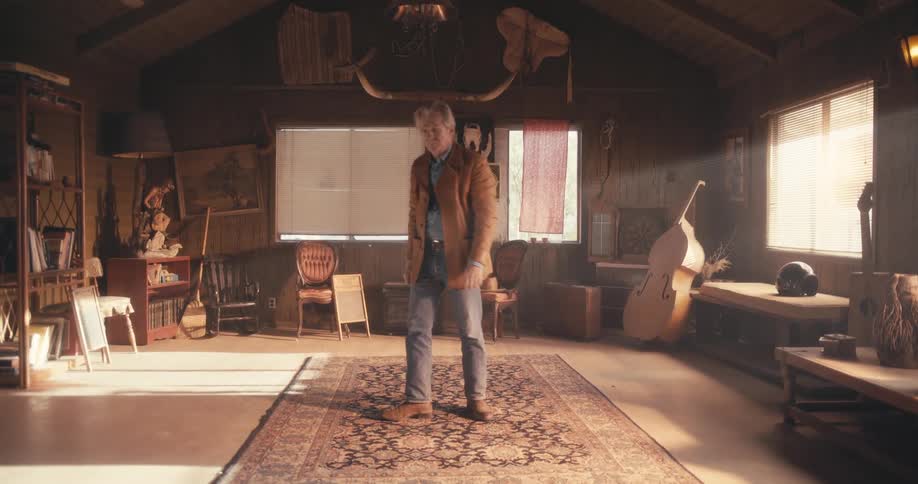 An old man standing in an old fashioned house checks his watch before gazing into the camera and walking away
