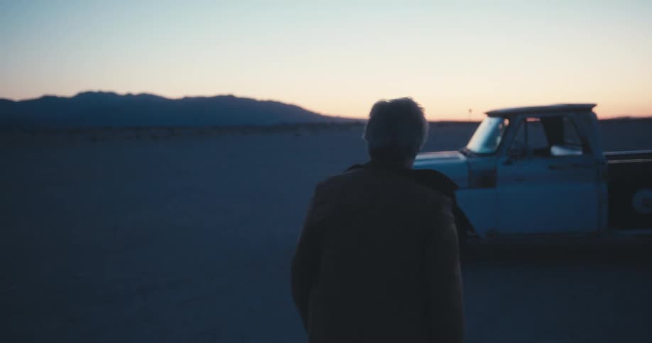 A man standing in a desert turns around and walks to his truck and gets in