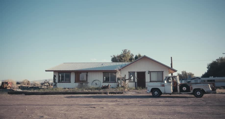 A man pulls his truck up to a house in a rural area and shields his eyes from the sun as he walks towards it