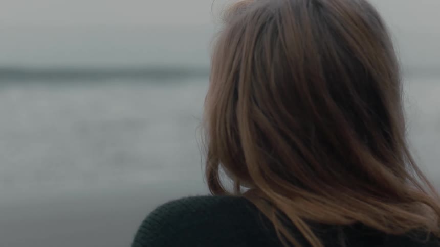 Blonde woman with a black sweatshirt watches the foamy waves breaking against the shore on the beach