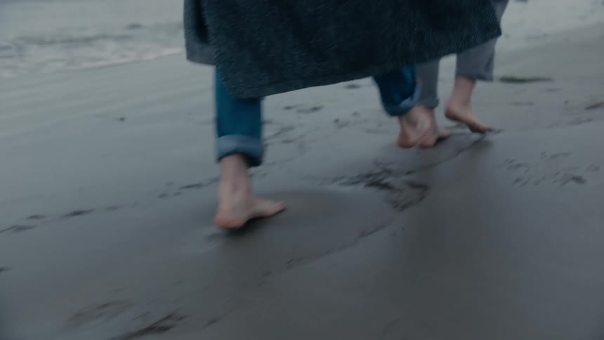 Two people walking on a sandy beach with one person trying to trip the other with the ocean the sky and clouds in the background