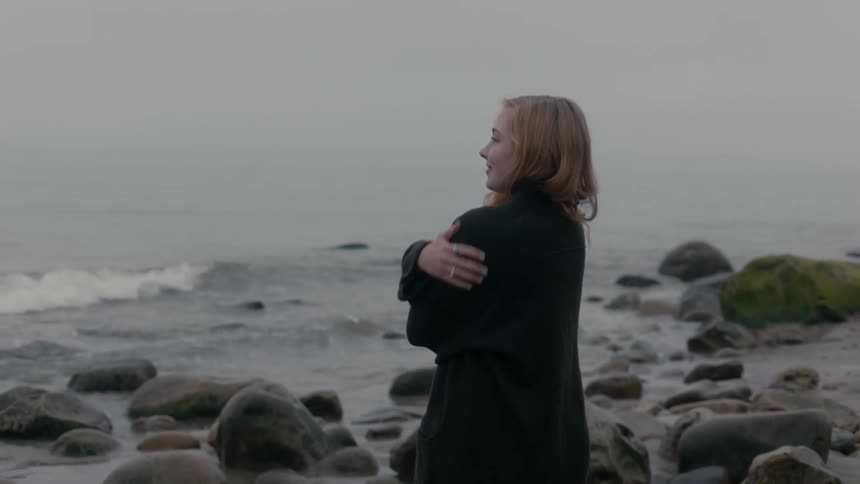A woman stands on a rocky beach wearing a sweater on a very cloudy day