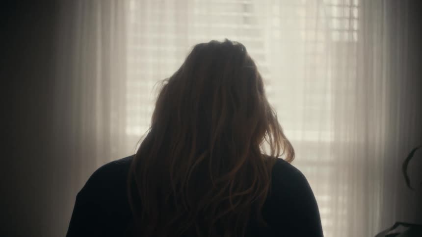 A woman is putting on a coat next to the window