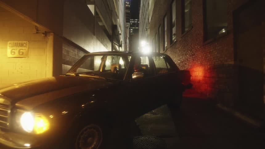 A man sits in a parked car in an alley way thats facing a parking garage