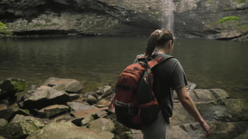 A woman carefully walks over rocks towards a waterfall feeding into a lake