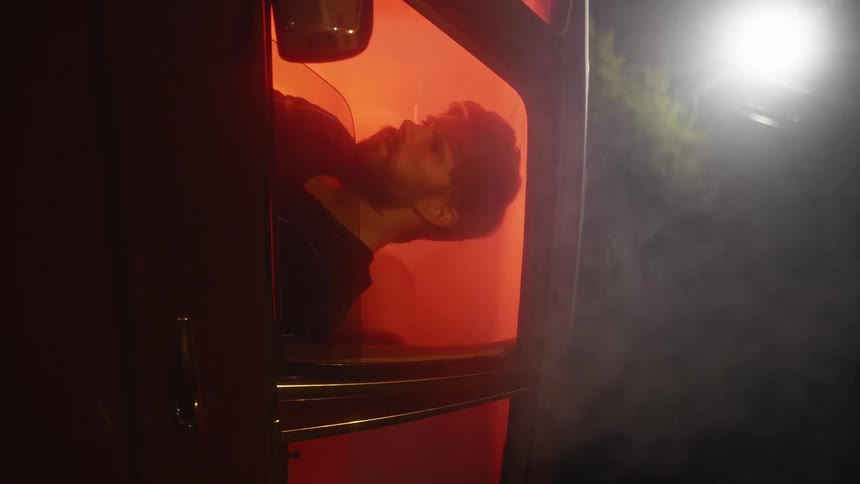 Man sits in drivers seat of smoking car lit internally by red light under streetlight and tree