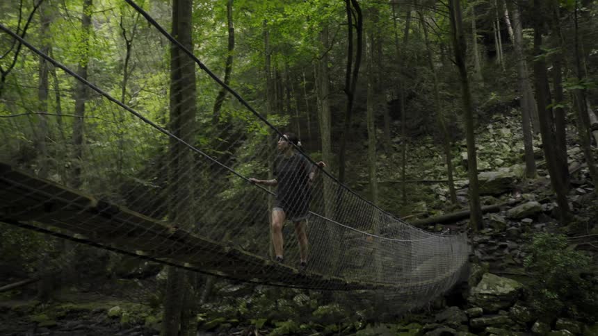 Woman walks across rope bridge leading from rocky tree strewn mountain slope over river with waterfall