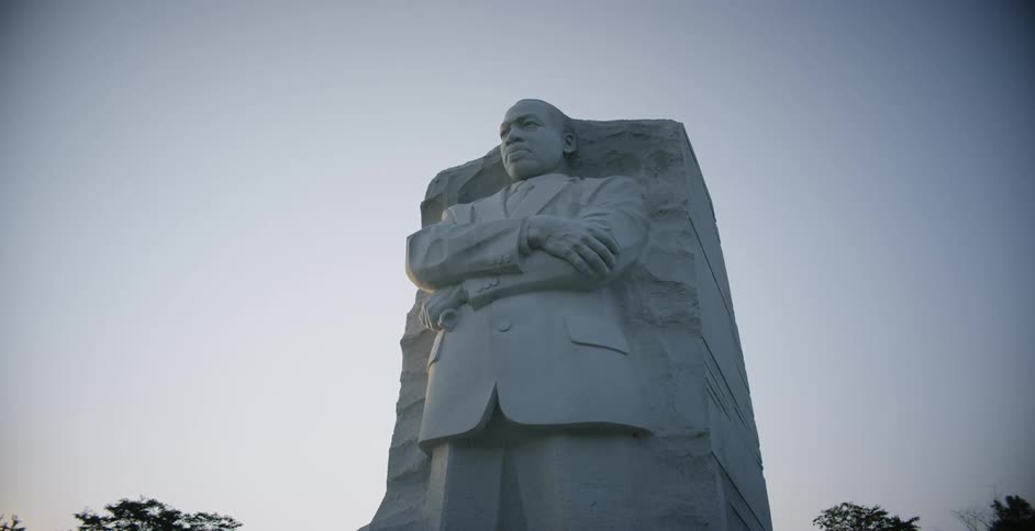 A large statue of a man in a suit with arms crossed carved partially out of stone at sunset with trees and the blue purple and pink sky in the background