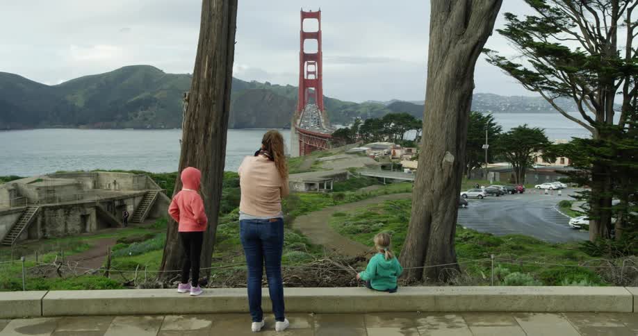 Mother and daughter stand while second daughter sits looking past park to ocean and bridge