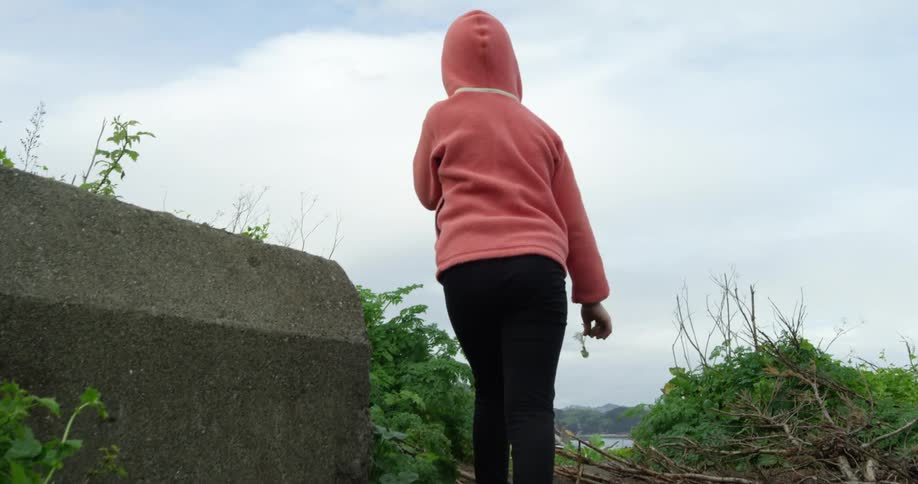 Girl in pink hoodie walks up incline until can view suspension bridge and opposite shore