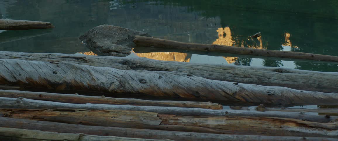 As water near long logs stops rippling the reflection of mountains becomes clearer