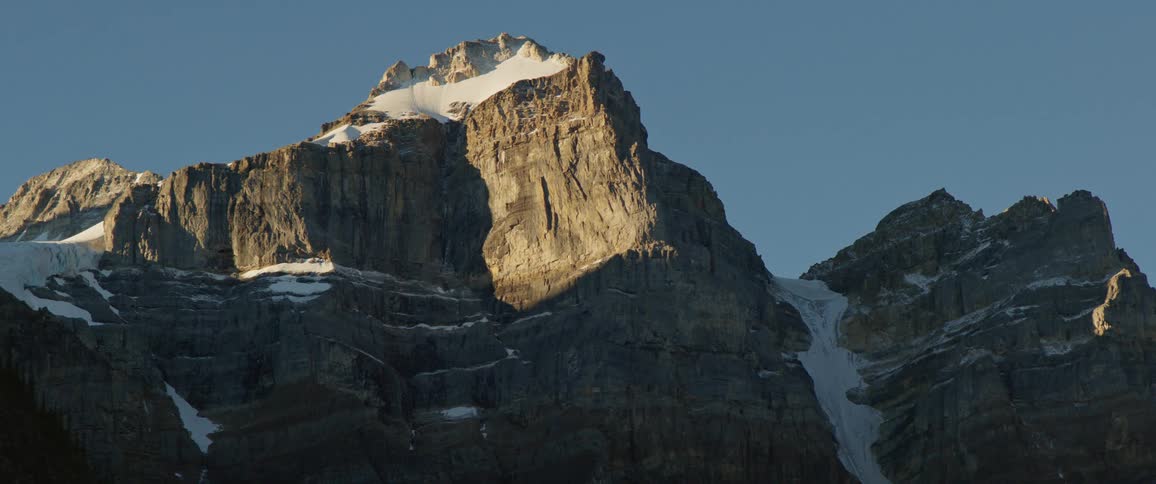 A stone mountain ridge dotted with patches of snow under a clear evening sky