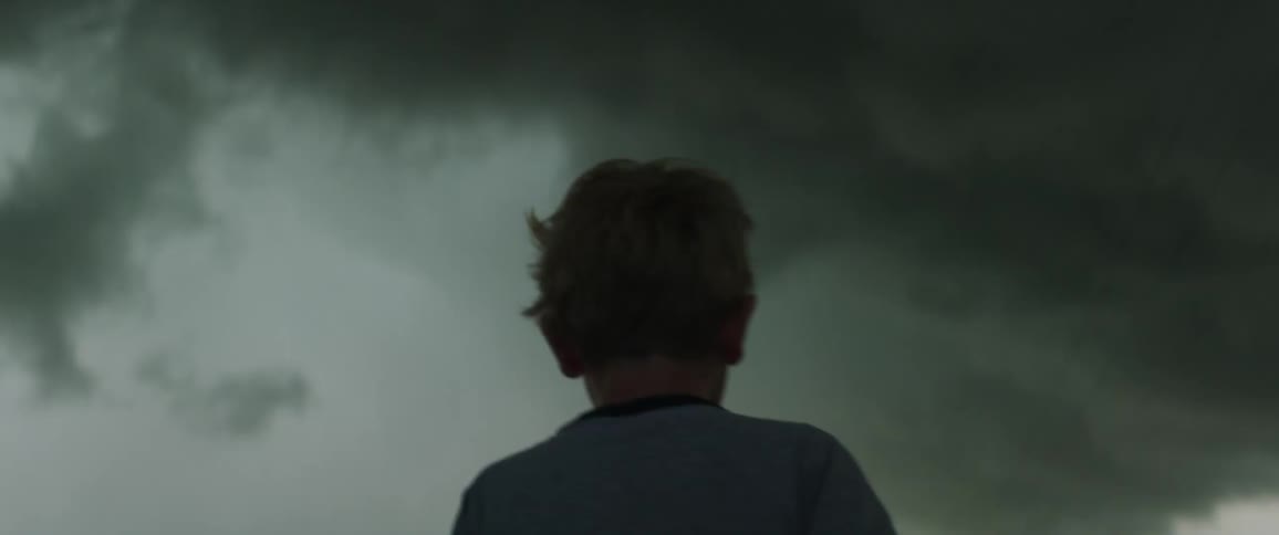 A boy walks a long a road through a field under a sky filled with thick dark clouds