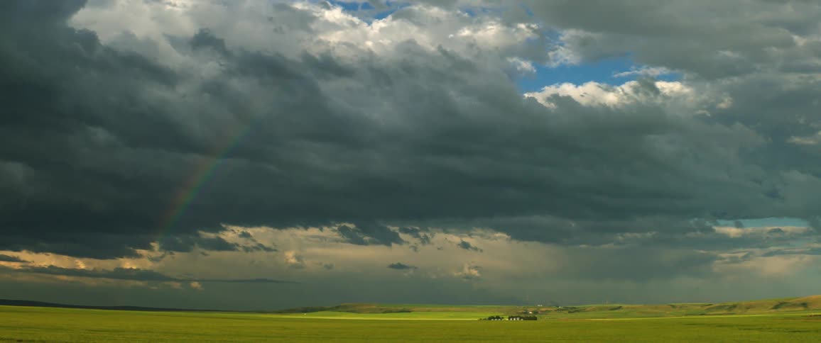 A mostly flat landscape with some hills has a visible rainbow caused by rain clouds and distant rain