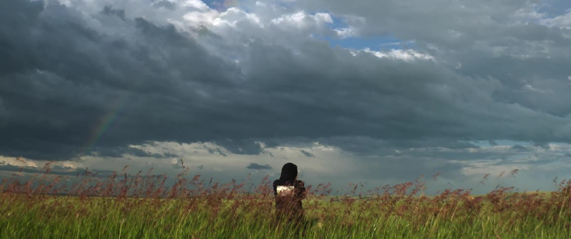 A person stands in the middle of a field taking a photo of the stormy sky