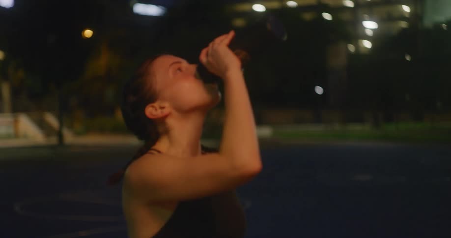 A sweaty woman in the park wearing a sports bra drinks from a bottle