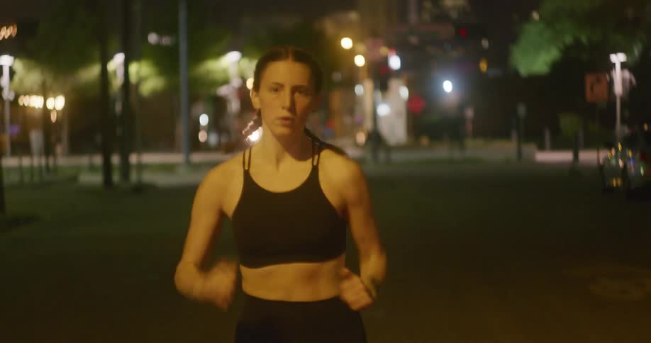 Woman wearing a black tank top and black pants running alone at night in front of flashing traffic lights trees and street lights past cars