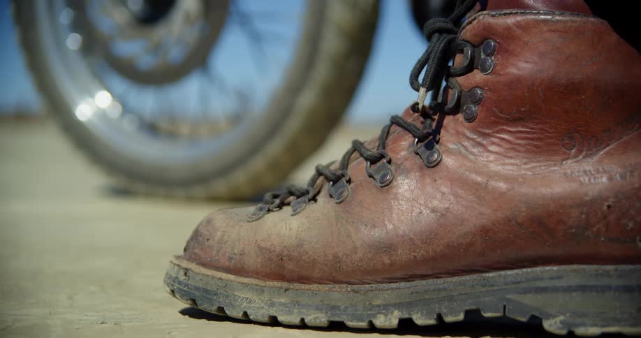 Close up of a red leather boot in front of the front wheel of a motorcycle outdoors