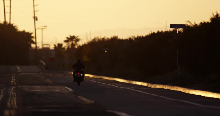 A silhouetted motorcycle driving on a paved road at sunset