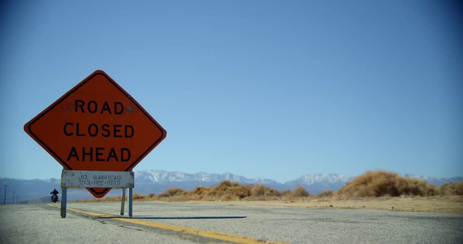 Motorcyclists rides past road sign ignoring road closure passing brown vegetation and mist covered mountains