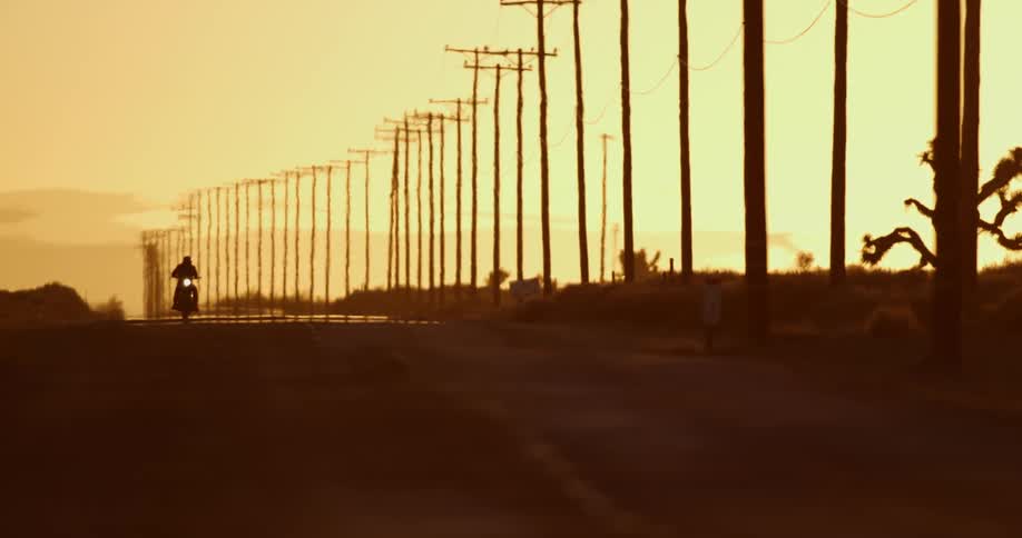 Motorcycle driving on a desert road next to a line of utility poles at sunset