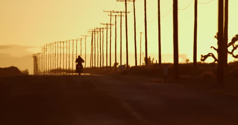 A motorcyclist riding down an empty road lined with telephone poles at dusk