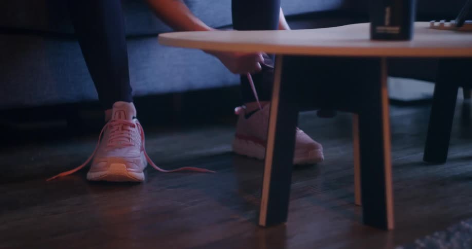 A woman takes off her pink sneakers while sitting on her living room couch