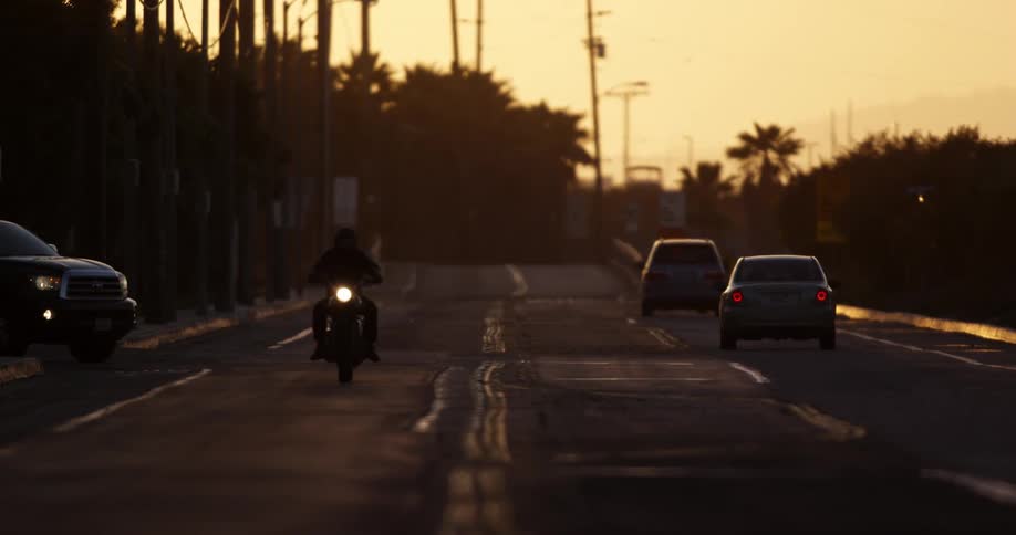 Cars and a motorcycle traveling on a paved road with an orange sky at sunset