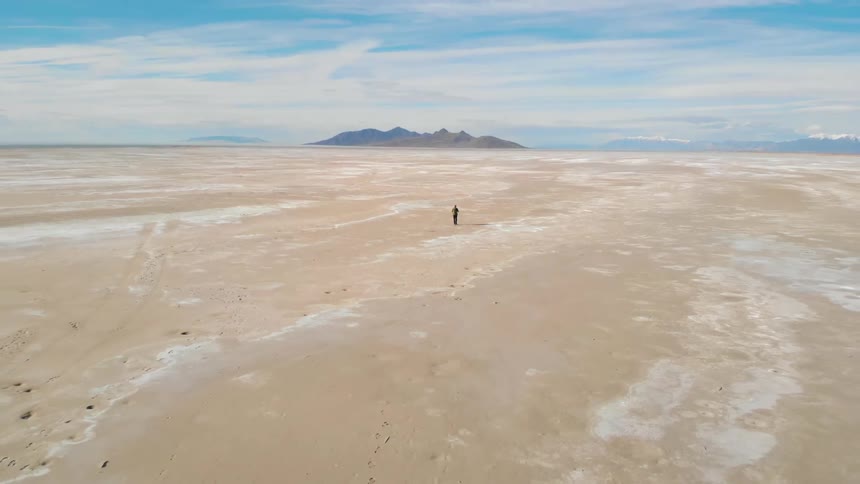 A person with a backpack walking in the desert with a mountain range the sky and clouds in the background