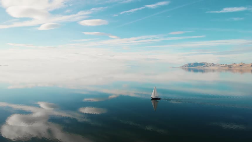 Sail boat is sailing across still water that is reflecting the bright blue sky and fluffy white clouds on a sunny day with islands in the distance