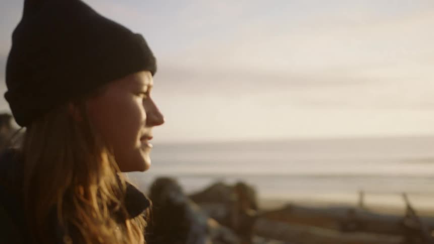 A young women walks along a shoreline in cold weather as she wears a backpack and a knit hat