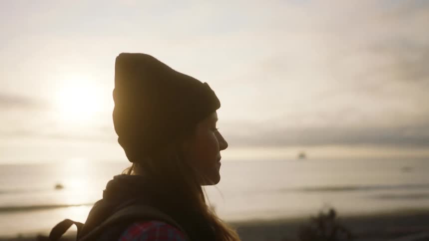 A woman wears a jacket and beanie walking along a rock filled beach also wearing a backpack