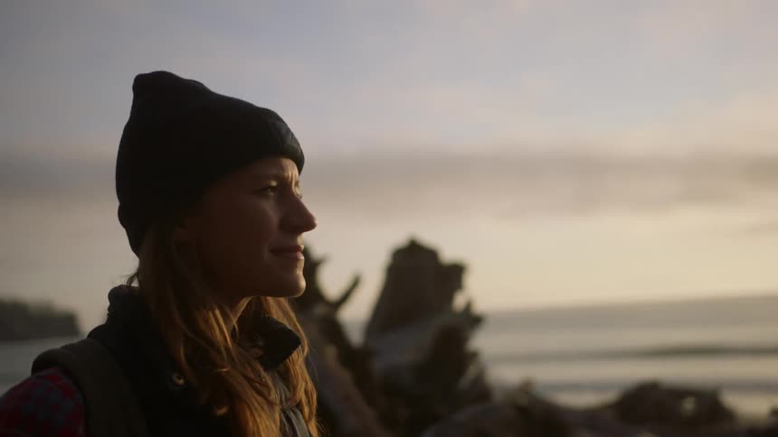 Woman with a beanie and brown hair looks around on the pebble beach at sunset